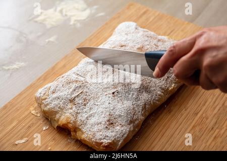 Hand mit einem Messer schneiden Bougaza, traditionelle Pudding Pie auf einem Holzbrett, Nahaufnahme. Griechisches Gebäck mit Sahnezucker-Pulver und gemahlenem Zimt Stockfoto