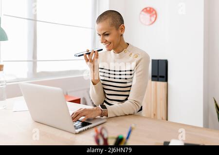 Trendige junge Geschäftsfrau, die Mobiltelefon benutzt, während sie zu Hause mit dem Computer arbeitet Stockfoto