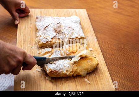 Hand mit einem Messer schneiden Bougaza, traditionelle Pudding Pie auf einem Holzbrett, Nahaufnahme. Griechisches Gebäck mit Sahnezucker-Pulver und gemahlenem Zimt Stockfoto
