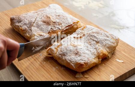 Griechisches Gebäck mit Sahnezucker-Pulver und gemahlenem Zimt. Hand mit einem Messer schneiden Bougaza, traditionelle Pudding Pie auf einem Holzbrett, Nahaufnahme Stockfoto