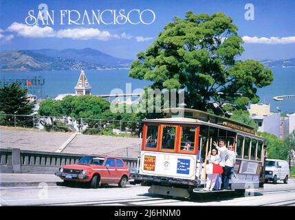 Eine Postkarte aus den 1980er Jahren, auf der Menschen an einem sonnigen Tag mit einer Seilbahn auf der Hyde Street in San Francisco, Kalifornien, USA, fahren. Der Turm im Hintergrund ist der Ghirardelli Building Tower und dahinter die San Francisco Bay Stockfoto