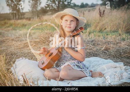 Porträt eines lächelnden Mädchens, das auf einer Decke im trockenen Heufeld sitzt, Picknick macht und das Spielen von Gitarren-Ukulele lernt. Ruhe und Entspannung. Korb in der Nähe Stockfoto