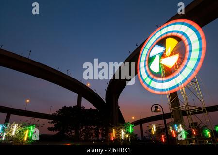 Ein farbenfroher Karneval- oder Themenpark unter einer Autobahnkreuzung, beleuchtete drehbare Turbinen und Neonlichter vor dem Sonnenuntergang. Stockfoto