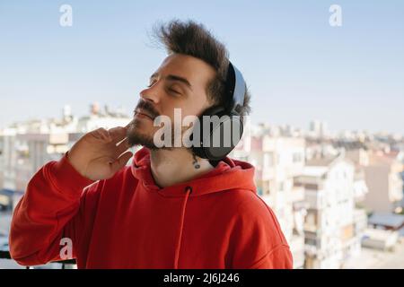 Kopfhörer, junger Mann, der Kopfhörer trägt und Musik auf dem Balkon hört, legte er seine Hand auf sein Kinn und ließ sich mit der Musik gehen Stockfoto