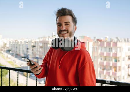 Posin, ein junger Gamer, der mit einem Telefon in der Hand für die Kamera posiert, nahm seine Kopfhörer ab und legte sie ihm auf den Hals, ein hübscher, erwachsener Mann, der lächelte Stockfoto