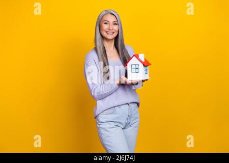 Foto von charmanten ziemlich pensionierten Frau tragen blauen Pullover mit kleinem Haus isoliert gelben Hintergrund Stockfoto