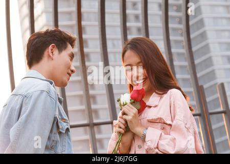 Junge glückliche Paare lieben und romantisch auf den ersten Tag Beziehung: asiatische Teenager-Frau überrascht und lächelt an Freund gibt rote Rosen-Blumen auf Dinne Stockfoto