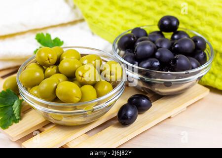 Viele grüne und schwarze marinierte ganze Oliven in der Glasschale auf hellem Holzbrett Hintergrund in der Küche. Stockfoto
