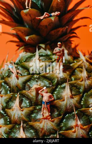 Nahaufnahme einiger Miniaturmenschen, die Badeanzug tragen, auf einer Ananas, vor einem orangen Hintergrund Stockfoto