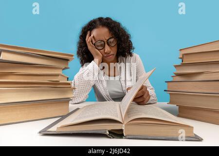 Müde junge schwarze Studentin, umgeben von Bücherstapeln, die Lehrbücher lesen, sich auf die Prüfung vorbereiten, sich gelangweilt fühlen Stockfoto