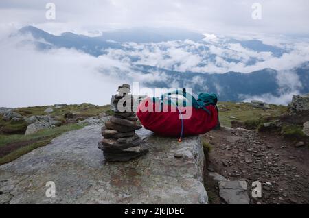 Touristischer Wanderrucksack auf Stein auf dem Berg Petros in den Karpaten. Steinpyramide im Vordergrund. Blick auf Wolken im Tal nach Regen Stockfoto