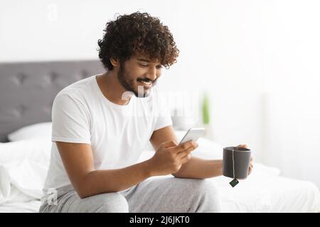 Fröhlicher indischer Mann trinkt Tee und benutzt Smartphone Stockfoto