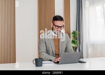 Selbstbewusster Geschäftsmann, der mit einem Laptop arbeitet. Fokussierter männlicher Unternehmer trägt Formalien. Er sitzt am Schreibtisch im Firmenbüro. Stockfoto