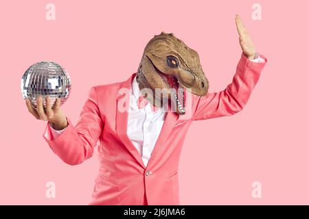 Modischer Mann in Dinosaurier-Gummimaske mit Disco-Ball in der Hand auf pastellrosa Hintergrund. Stockfoto