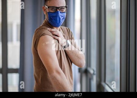 Porträt eines lächelnden geimpften Unternehmers während der Pandemie Covid19. Männlicher Profi, der Verbandszeug am Arm zeigt. Er trägt eine Brille und schützt Stockfoto