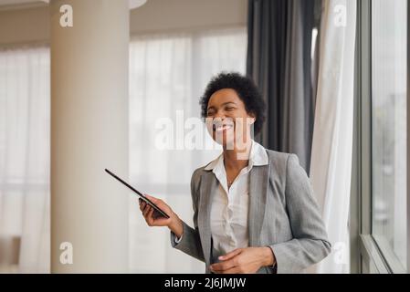 Fröhliche Geschäftsfrau mit digitalem Tablet. Glückliche weibliche Profi trägt formals. Sie steht mit geschlossenen Augen im Büro. Stockfoto