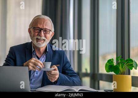 Lächelnder Geschäftsmann beim Kaffee. Porträt von älteren glücklichen Profi arbeiten auf Laptop. Er sitzt am Schreibtisch im Büro Stockfoto