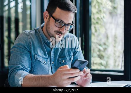 Junge männliche Unternehmen professionelle SMS auf Smartphone. Geschäftsmann trägt Denim-Hemd. Er arbeitet am Tisch im Heimbüro Stockfoto