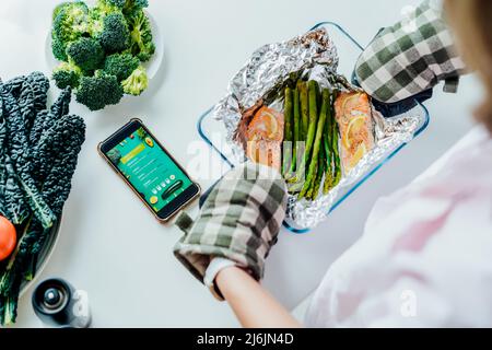 Pescetarian Mahlzeit Diät Rezepte. Draufsicht Frau in Form mit gebackenem Lachs und Spargel. Telefon mit Pescetarian Menü im Handy liegt auf dem Tisch. Stockfoto