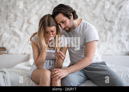 Verärgerte junge Frau mit negativem Schwangerschaftstest weinend auf dem Bett, liebevoller Ehemann, der sie zu Hause unterstützt Stockfoto