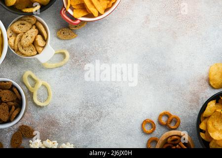 Snacks. Ungesunde Lebensmittel. Alle klassischen Kartoffelsnacks mit Erdnüssen, Popcorn- und Zwiebelringen und gesalzenen Brezeln in Schüsseln auf grauem Beton-Hintergrund Stockfoto