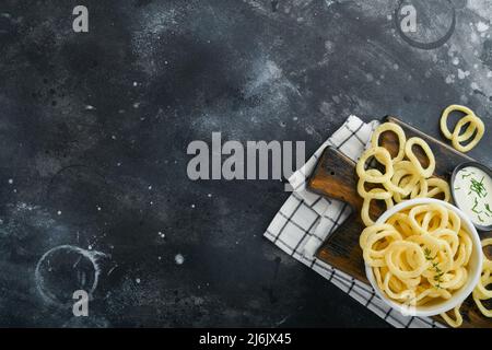 Zwiebelringe. Fast Food. Knusprige Zwiebelchips in schwarzer Keramikschüssel mit saurer Rahmsoße und Zwiebeln in Holzständer auf altem schwarzen Betonhintergrund. Am Stockfoto