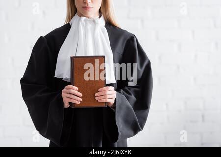 Teilansicht des Staatsanwalts in schwarzem Mantel hält Buch in der Nähe der weißen Wand Stockfoto