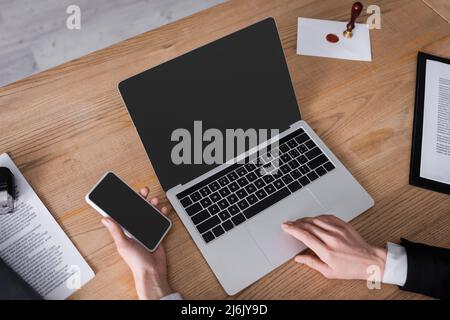 Teilweise Ansicht des Anwalts, der das Mobiltelefon in der Nähe des Laptops mit leerem Bildschirm und Dokumenten hält Stockfoto