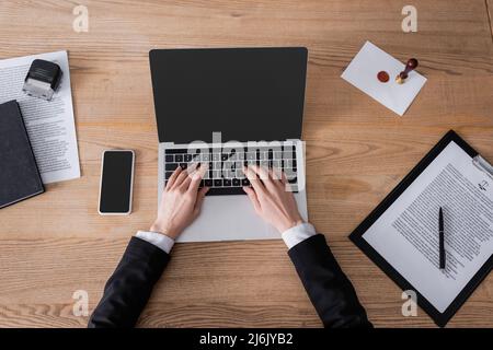 Draufsicht auf einen zugeschnittenen Anwalt, der in der Nähe des Smartphones auf dem Laptop tippt, mit leerem Bildschirm und Vertrag Stockfoto