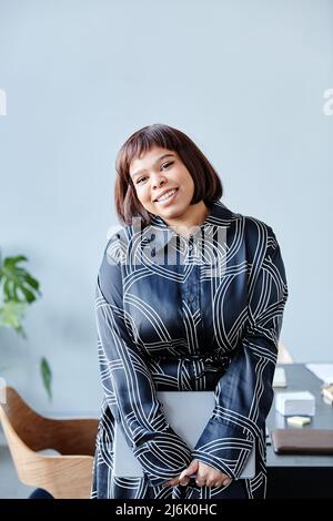 Minimalistisches Porträt einer jungen schwarzen Frau mit kurzem Haarschnitt, die fröhlich auf die Kamera lächelt, während sie im Büro gegen Blau steht Stockfoto