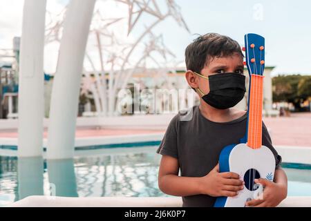 Lateinischer Junge im Freien mit einer medizinischen Maske und einer Ukulele in den Händen. Konzepte der musikalischen Ausbildung Stockfoto