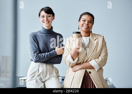 Auffältige Porträt von zwei jungen Geschäftsfrauen, die vor einfachem Hintergrund vor der Kamera im Büro lächeln Stockfoto