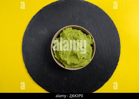 Keramikschale gefüllt mit der leckeren Sauce aus Avocado, Knoblauch, Limette und Tomaten, die als Guacamole bekannt sind, auf einem Schieferteller auf gelbem Grund Stockfoto