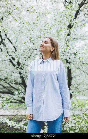 Junge hübsche Frau genießt es, in der Nähe des blühenden Frühlingsbaums zu stehen. Ein Mädchen mit beigefarbenem Hut und weißem Kleid lächelt zwischen blühenden Apfelbäumen. Frühlingsmeere Stockfoto