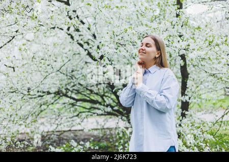 Junge hübsche Frau genießt es, in der Nähe des blühenden Frühlingsbaums zu stehen. Ein Mädchen mit beigefarbenem Hut und weißem Kleid lächelt zwischen blühenden Apfelbäumen. Frühlingsmeere Stockfoto