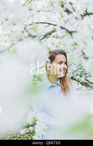 Junge hübsche Frau genießt es, in der Nähe des blühenden Frühlingsbaums zu stehen. Ein Mädchen mit beigefarbenem Hut und weißem Kleid lächelt zwischen blühenden Apfelbäumen. Frühlingsmeere Stockfoto