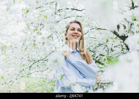 Junge hübsche Frau genießt es, in der Nähe des blühenden Frühlingsbaums zu stehen. Ein Mädchen mit beigefarbenem Hut und weißem Kleid lächelt zwischen blühenden Apfelbäumen. Frühlingsmeere Stockfoto