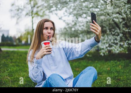 Draufsicht auf die Frau, die im Parkgarten auf dem grünen Gras mit Smartphone, Kopfhörer, Tablet, Buch und Kaffee in der Hand sitzt Stockfoto