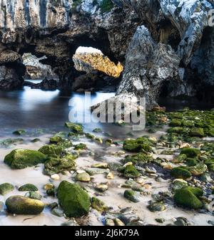 Strand mit Klippen und Höhlen und algenbedeckten Felsen auf goldenem Sand Stockfoto