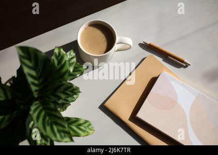 Ästhetischer femininer Arbeitsplatz. Tasse Kaffee, Notizbücher, grüne Pflanze auf dem Schreibtisch des Heimbüros bei Sonnenuntergang. Stockfoto