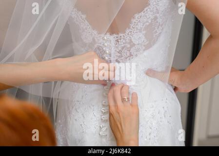 Die Brautjungfern helfen der Braut, ihr Hochzeitskleid anzuziehen. Nahaufnahme der Hände Stockfoto