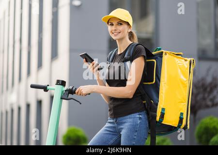 Low-Angle-Ansicht der Zustellfrau mit Thermo-Rucksack über die Navigations-App auf dem Mobiltelefon. Weibliche Kurierin auf der Suche nach Kundenadresse mit Smartphone Reiten Auftrag auf Elektroroller. Stockfoto