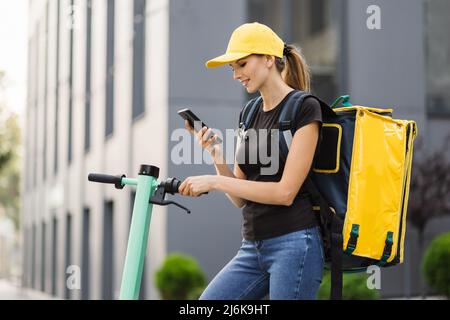 Low-Angle-Ansicht der Zustellfrau mit Thermo-Rucksack über die Navigations-App auf dem Mobiltelefon. Weibliche Kurierin auf der Suche nach Kundenadresse mit Smartphone Reiten Auftrag auf Elektroroller. Stockfoto
