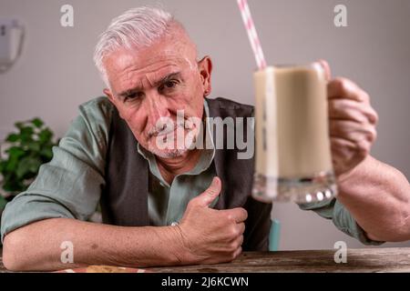 Lässig gekleideter reifer Mann mit Sonnenbrille sitzt an einem Tisch und trinkt Eiskaffee durch einen Strohhalm Stockfoto