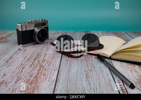 Kamera, Sonnenbrille mit einem Notizbuch und einem Stift auf einem Holztisch, auf grünem Hintergrund. Urlaubskonzept. Stockfoto