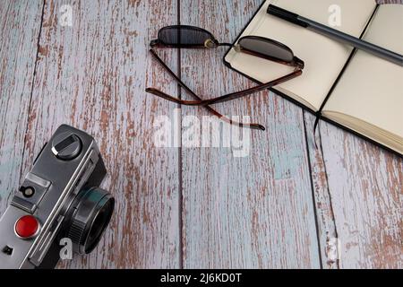 Blick von oben auf ein Notizbuch, eine Sonnenbrille, eine Fotokamera und einen Stift auf einem weißen Holztisch. Stockfoto