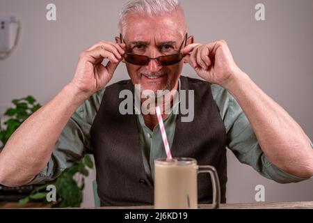 Lässig gekleideter reifer Mann mit Sonnenbrille sitzt an einem Tisch und trinkt Eiskaffee durch einen Strohhalm Stockfoto