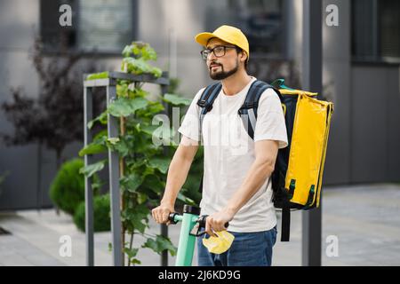 Fahrer bärtigen Mann in gelbe Kappe und thermische Box Rucksack liefern Mahlzeit an Kunden mit Elektroroller. Ökologisches Fast-Delivery-Food-Konzept. Stockfoto