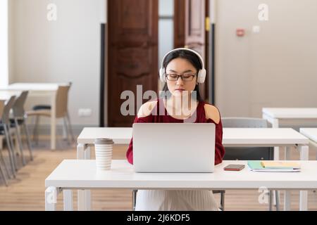 Serious Asian Student Mädchen in Kopfhörern beobachten pädagogische via Internet auf Laptop in der Bibliothek Stockfoto