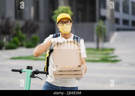 Lieferkurier, der die Pizza in Kartons anbietet. Delivery man auf medizinische Maske und weiße Handschuhe mit Pizza-Box aus der Nähe. Lieferung männlich hält einen Stapel von Pizzakartons. Pizzalieferung. Stockfoto